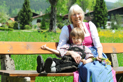 lächelnde Oma sitzt mit Enkel auf einer Parkbank, Oma hält ihren Enkel mit dem rechten Arm umschlungen, in ihrer linken Hand hält sie einen selbst gepflückten Wiesenstrauß